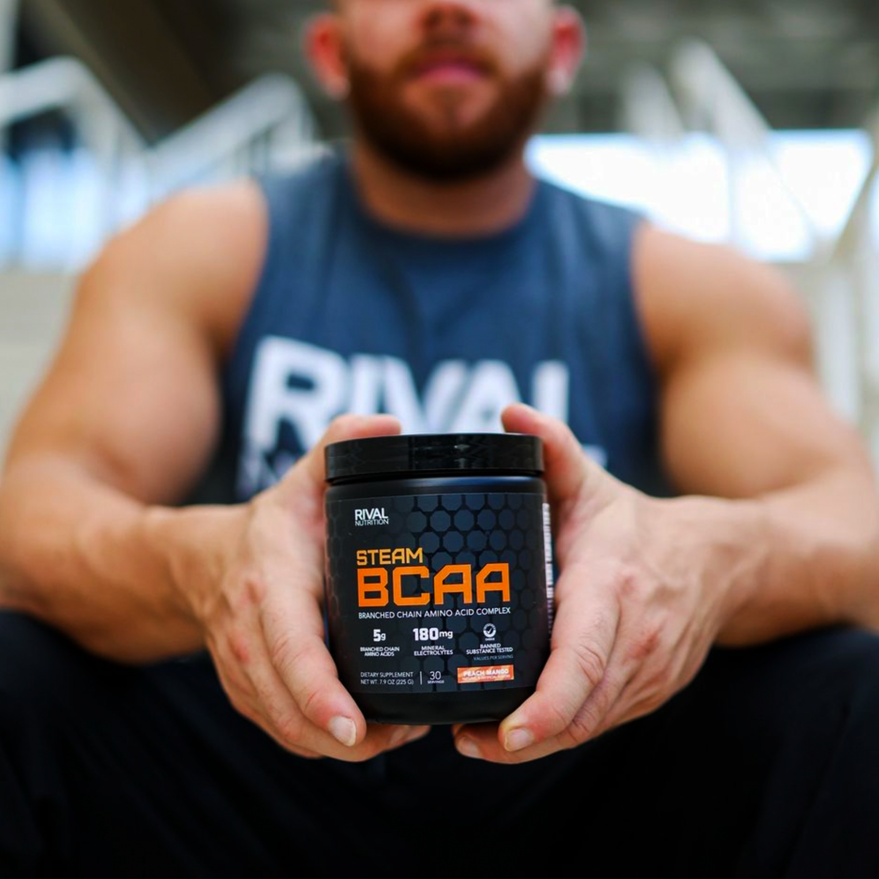 A fit Rival Nutrition Athlete wearing a cut off Grey Short Sleeve Rival Nutrition Shirt and holding a bottle of Rival Nutrition Steam BCAA Branched Chain Amino Acid Complex Peach Mango Flavor to the camera.